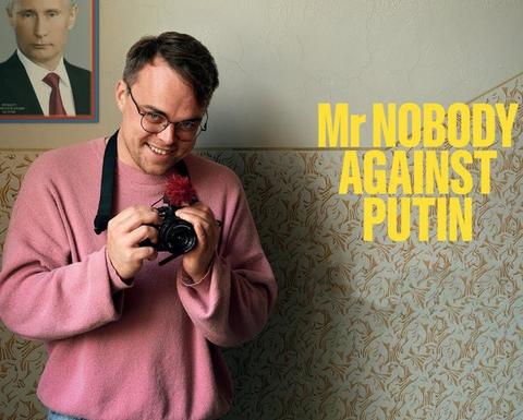 Film poster for Mr Nobody Against Putin. A smiling man in glasses and a pink sweater holds a camera with a red fuzzy microphone. Behind him is patterned wallpaper and a framed portrait of Vladimir Putin. The film’s title, “Mr Nobody Against Putin,” appears in large yellow letters on the right.